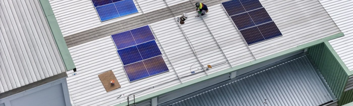 technician in safety gear inspects solar panels on a metal rooftop, holding a tool. The scene emphasizes renewable energy, solar technology, and maintenance of sustainable infrastructure.